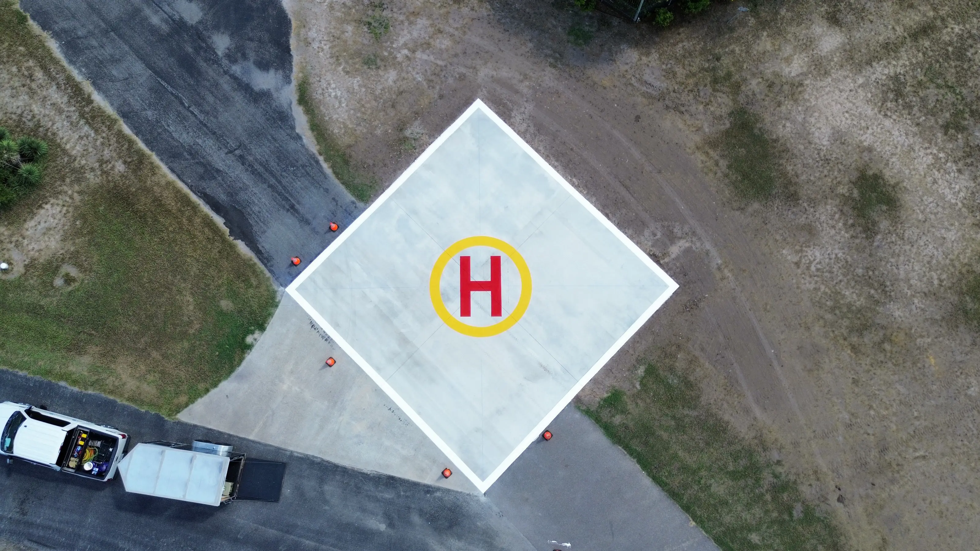 Wide aerial drone view of a freshly painted helipad — white pad, yellow circle, and red H — with the Coastal Roadworks truck and trailer nearby