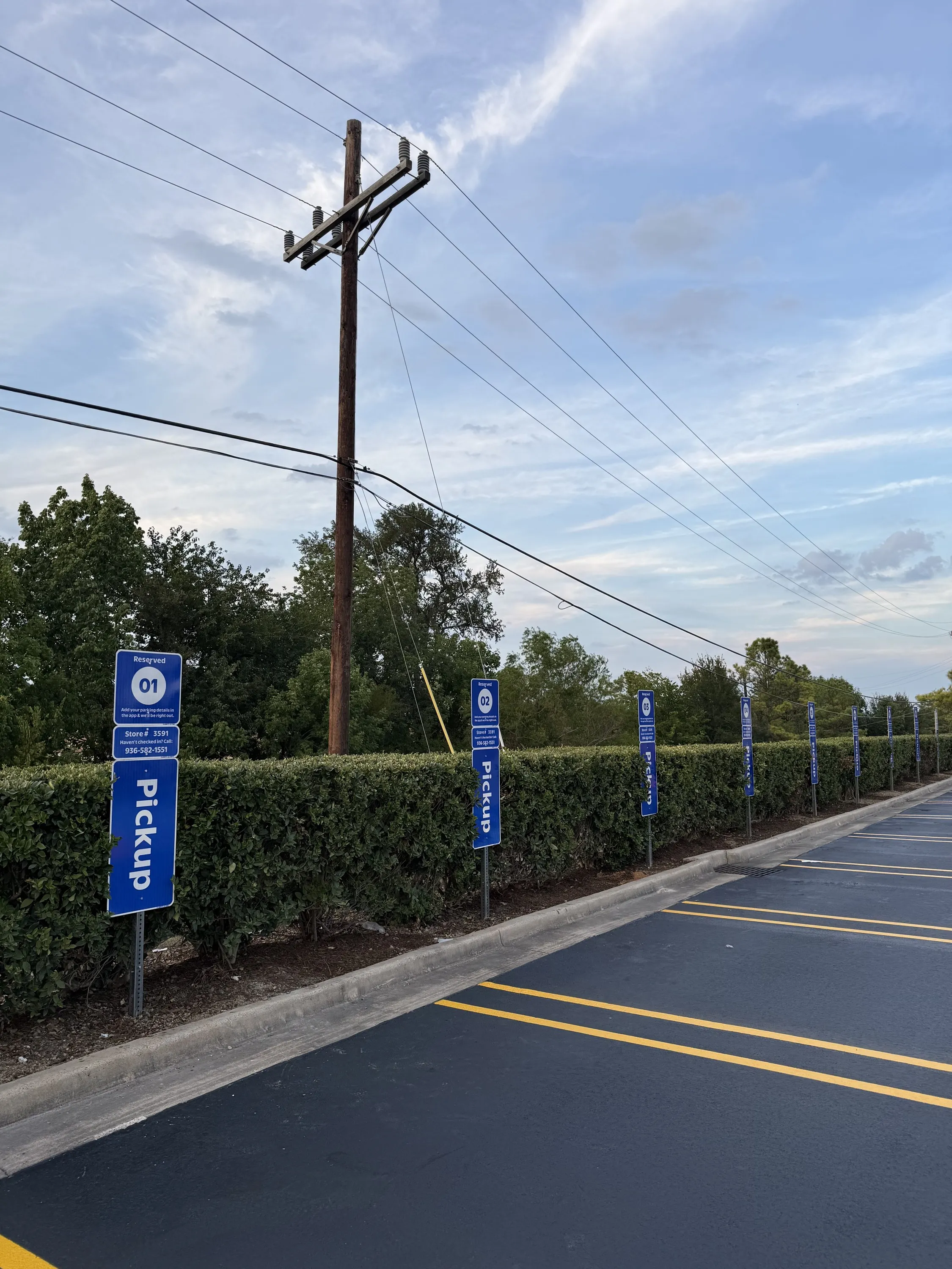 Row of blue bollard-mounted pickup signs along a hedge beside a freshly sealed and striped lot