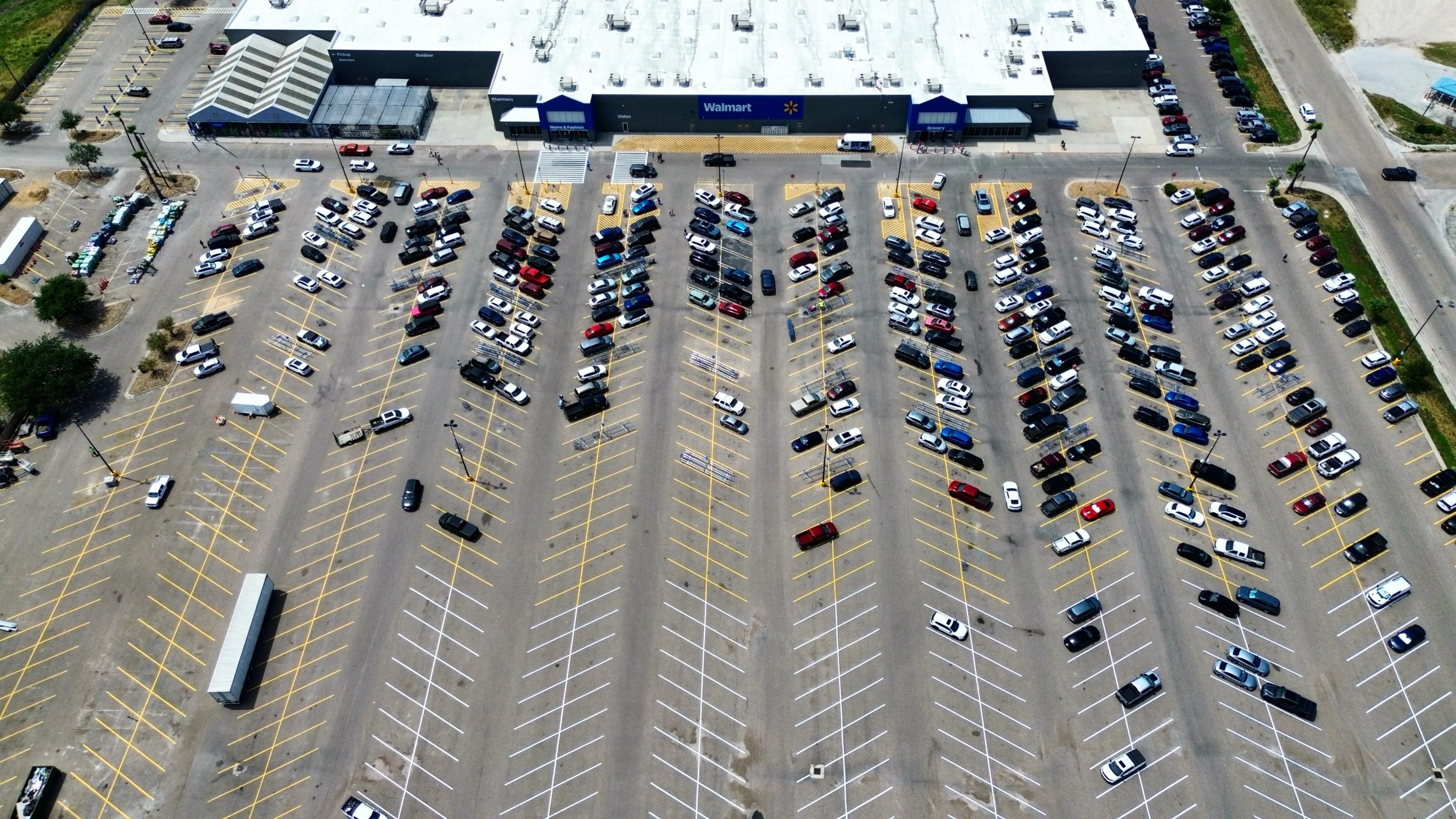 Aerial view of a large asphalt retail parking lot in daily use — the kind of surface that takes pothole and patch work