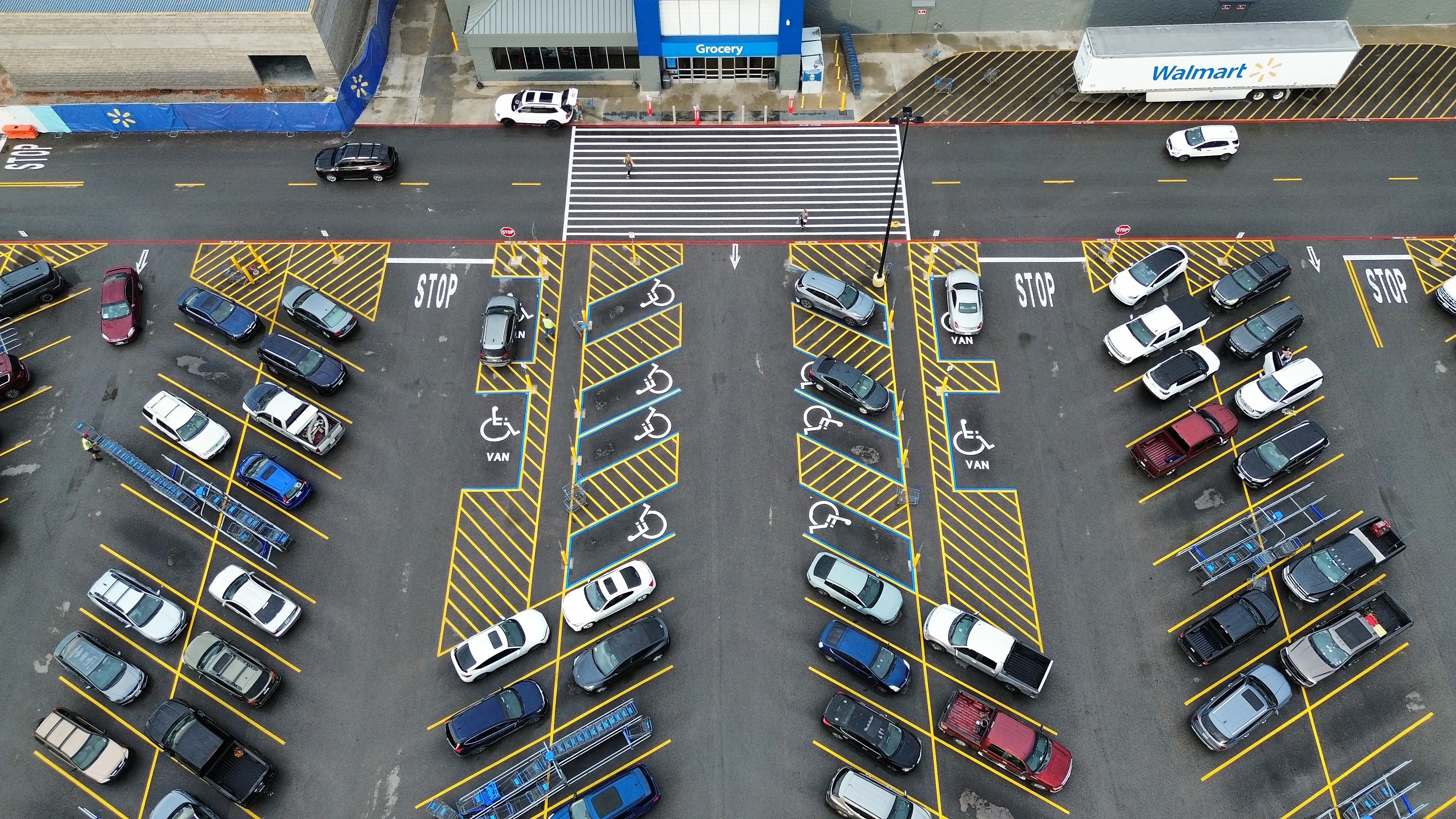 Top-down aerial of a dark sealed supercenter lot with fresh yellow stalls, accessible symbols, and STOP stencils